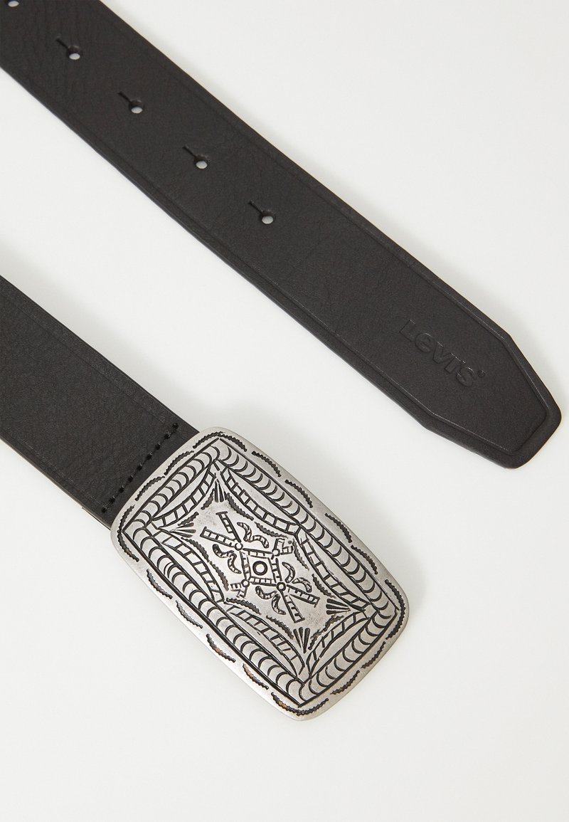 Ceinture en cuir noir avec une large boucle métallique argentée présentant des motifs en relief complexes et une forme rectangulaire.