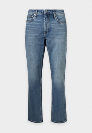 Jeune homme debout face à l'avant, portant un t-shirt blanc uni, un jean bleu et des chaussures noires élégantes, une main dans la poche.