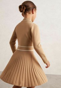 Kootud kleit beežis, millel on istuv ülemine osa ja volditud seelik. Vöö vahel on valge aksent ja pika varrukaga, esitledes tekstuuri ja disaini.