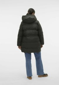 Manteau doudoune vert foncé avec capuche, design matelassé, taille cintrée et coupe légèrement oversize, associé à un jean bleu évasé et des chaussures noires.