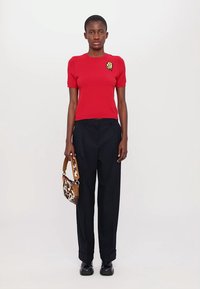 Top corto a maniche corte rosso in maglia con una toppa sul petto; abbinato a pantaloni scuri a vita alta e una borsa a tracolla marrone e bianca a fantasia.