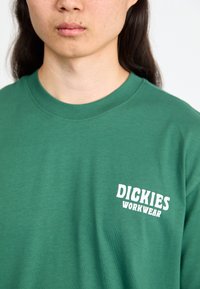 Zelena pamučna majica s okruglim dekolteom i kratkim rukavima, na lijevoj strani prsnog dijela otisnuto "DICKIES WORKWEAR" u bijeloj boji.