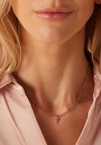 Zilveren ketting met verlengde schakels en een klein kruisje als hanger, gedragen over een lichtroze blouse, wat het elegante ontwerp accentueert.
