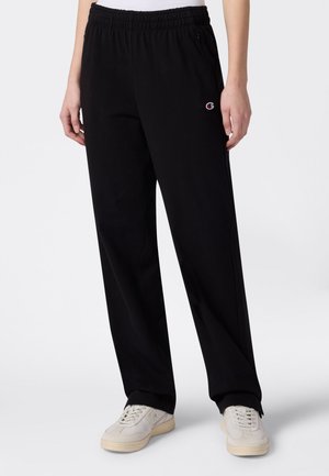 Pantaloni della tuta neri a vestibilità comoda con elastico in vita e tasca con cerniera, indossati con sneakers color avorio su sfondo bianco.