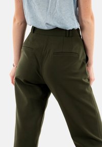 Olivově zelené kalhoty na míru s hladkou texturou, vybavené dvěma zadními kapsami a vysokým pasem, stylizované s světle šedým topem.