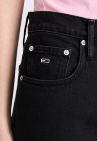 Jeans de mezclilla negros con herrajes plateados, que presentan un pequeño logo bordado en el bolsillo delantero. La tela tiene una textura suave.