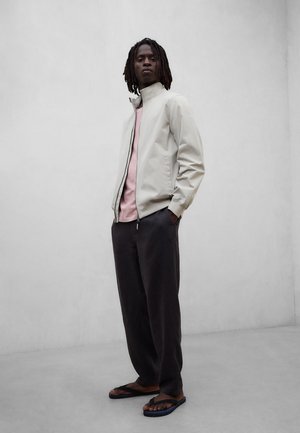 Muž s dredy, oblečený v béžové bundě, růžové košili, tmavých volných kalhotách a černých žabkách, stojící před jednoduchým světle šedým pozadím.