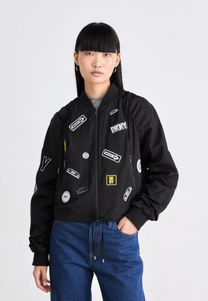 Jachetă bomber neagră cu multiple petice din diferite țesături și logo-uri, inclusiv DKNY. Prezintă un sistem de închidere cu fermoar și manșete elastice, purtată cu blugi blue.