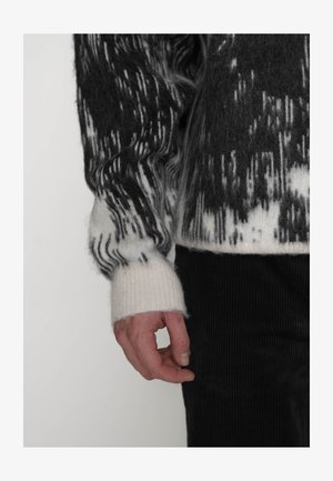 Mano appoggiata accanto al corpo che indossa un maglione peloso a motivo bianco e nero con polsino a coste e pantaloni di velluto a coste neri.