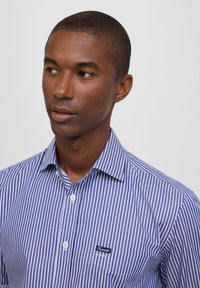Camicia a righe blu e bianche, con design a bottoni, dotata di una piccola tasca sul petto e logo. Tessuto liscio con colletto classico.