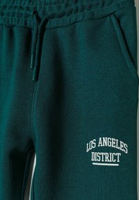 Pantaloni della tuta color teal scuro con una consistenza morbida, caratterizzati da un logo bianco "LOS ANGELES DISTRICT" e una vita elasticizzata con laccetti regolabili.