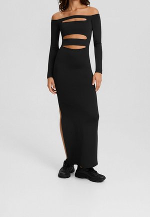 Frau trägt ein langes schwarzes schulterfreies Kleid mit Ausschnitten an Brust und Taille, kombiniert mit schwarzen klobigen Sneakers.