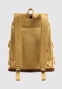 Gele canvas rugzak met twee brede banden, een handvat aan de bovenkant en een bruine onderrand. Bevat zijzakken en heeft een gestructureerde vorm.