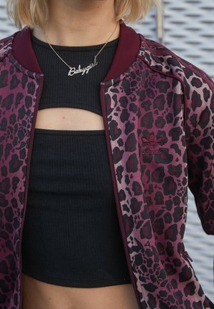 Close-up van een persoon die een bordeauxrode en zwarte Adidas jas met luipaardprint draagt over een zwarte crop top met een "Babygirl" ketting.