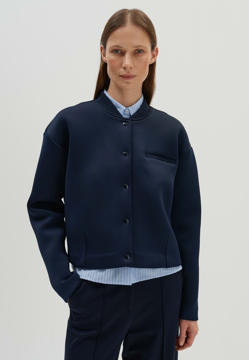 Marineblaue Jacke mit rundem Kragen und Knopfverschluss, mit einer Brusttasche, aus glattem Stoff, getragen über einem hellblauen gestreiften Hemd.