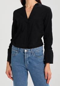 Blouse noire à manches longues avec un col en V et des manches froncées. Associée à un jean bleu clair taille haute. Le tissu semble lisse et léger.