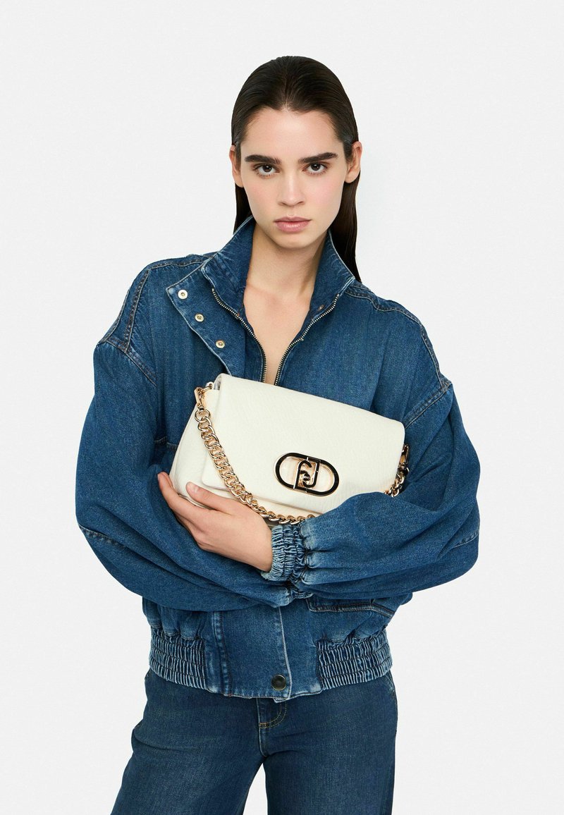 Borsa in pelle bianca con tracolla a catena dorata e chiusura ovale, appoggiata su una giacca di denim blu. Maniche a campana e vita aderente.
