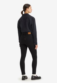 Žena s copom, oblečená v čiernej košeli s gombíkmi, čiernych vysokých slim džínsoch, bielych ponožkách a čiernych mokasínach, stojí postavená bokom.