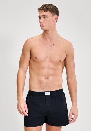 Joven sin camiseta con cabello corto y rizado, vistiendo pantalones cortos negros con cintura elástica, mirando hacia su izquierda contra un fondo claro y liso.