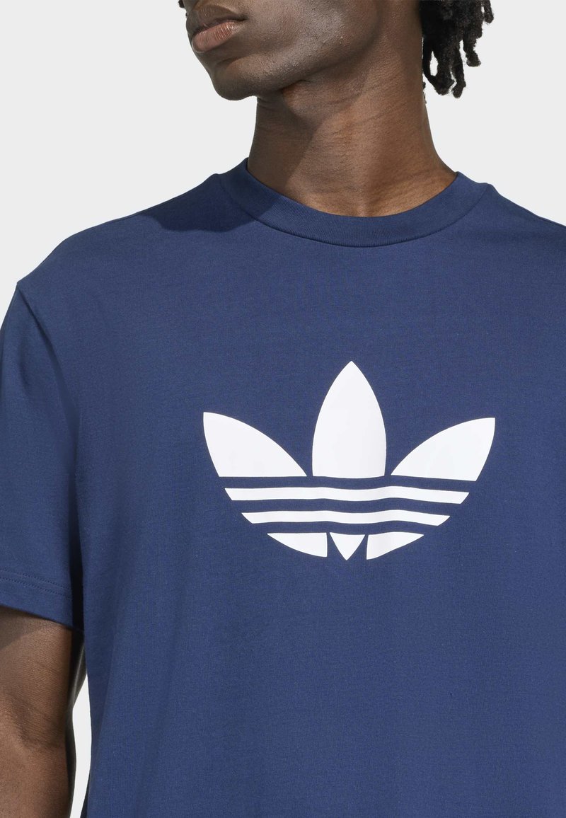 Uomo che indossa una maglietta blu navy con grande logo bianco Adidas trefoil centrato sul petto, rivolto leggermente a sinistra.
