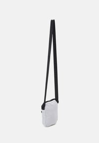 Gray crossbody bag with a black adjustable strap, rectangular shape, zip closure, and subtle logo detailing on the strap. Lightweight material.
