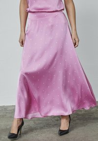 Lyserød satin maxi-nederdel med hvide polka dutter, flydende design, glat tekstur og en subtil glans. Kombineret med sorte højhælede sko.