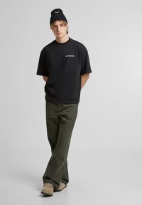Zwarte t-shirt met korte mouwen en witte tekst, olijfgroene broek, beige schoenen en een zwart gebreide beanie met een logo. Eenvoudige, casual outfit.
