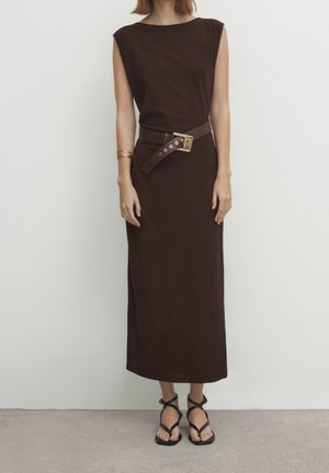 Robe sans manches marron en tissu doux, dotée d'une ceinture avec une boucle dorée, associée à des sandales noires à lanières. Fond uni.