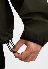 Mörkgrön jackärm med elastisk trim. En hand drar i en svart dragsko, vilket avslöjar texturerad tyg och en tätt sittande design.