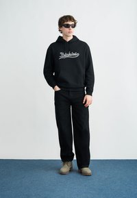 Μαύρο φούτερ με κουκούλα και λογότυπο "Dickies", συνδυασμένο με μαύρο παντελόνι και μπεζ παπούτσια, τοποθετημένο σε ανοιχτό φόντο με μπλε χαλί.