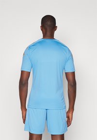 Camisa atlética azul clara de mangas curtas com calções combinando. Apresenta detalhes dourados nos ombros. Textura suave, design desportivo.