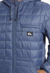 Blaue gepolsterte Jacke aus glänzendem Material, mit Reißverschluss vorne, einer Brusttasche und einem Logo-Patch auf der linken Seite.