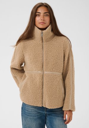 Junge Frau mit langen braunen Haaren, die eine beige Fleecejacke mit Reißverschluss und blaue Jeans trägt, steht vor einem schlichten hellen Hintergrund.