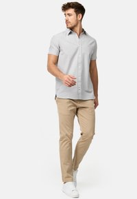 Camisa de botones de manga corta en gris con cuello, combinada con pantalones beiges. Modelo vistiendo zapatillas blancas, de pie sobre un fondo liso.