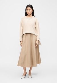 Γυναίκα με μακριά σκούρα μαλλιά, φορόντας μπλούζα μπεζ με μακριά μανίκια, τζίνι midi σε ίδιο χρώμα και λευκά μυτερά τακούνια, στέκεται μπροστά σε απλό φόντο.