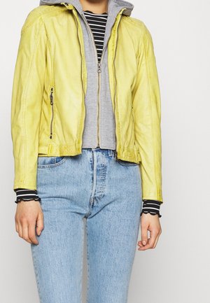 Persona che indossa una giacca di pelle gialla sopra una felpa con cappuccio grigia con zip e una maglietta a righe bianca e nera, abbinata a jeans azzurri.