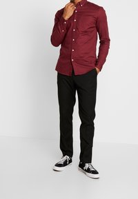 Muž v zapnuté bordó košili, černých kalhotách a černých teniskách s bílými tkaničkami, stojící na pozadí z čistě bílé barvy.