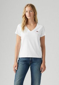 Camiseta blanca de cuello en V hecha de algodón, con mangas cortas y un pequeño logotipo rojo de Levi's en el lado izquierdo del pecho. Lleva puestas jeans azules.