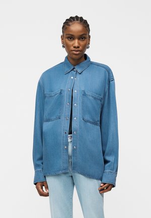 Młoda osoba z warkoczami nosi oversize'ową niebieską dżinsową koszulę z dwoma kieszeniami na piersi, jasnoniebieskie dżinsy i srebrne kolczyki koła.