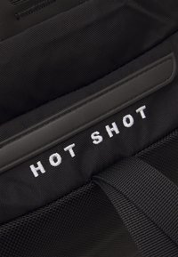Tissu noir avec surface texturée comportant un texte brodé blanc "HOT SHOT" et une sangle noire attachée.
