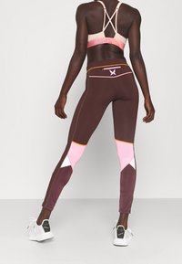 Leggings marroni con pannelli rosa e bianchi, vita alta, cuciture a contrasto e una tasca con cerniera. Abbinati a un reggiseno sportivo rosa con bretelle.