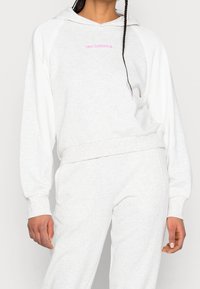 Jasnoszara bluza z białymi rękawami raglanowymi, z różowym logo "new balance" na piersi. Miękka tekstura; noszona z dopasowanymi spodniami dresowymi.