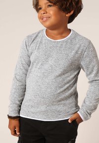 Grijze lange mouwen shirt van zacht, gestructureerd stof met een witte ronde hals en contrasterende stiksels. Gecombineerd met zwarte broeken en een polsaccessoire.