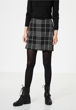 Femme portant une jupe à carreaux noire et blanche, des collants noirs, des bottines noires lacées et un haut noir à manches longues, debout sur un fond blanc.