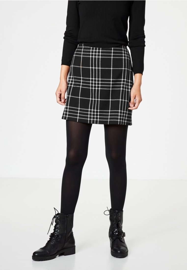 Femme portant une jupe à carreaux noire et blanche, des collants noirs, des bottines noires lacées et un haut noir à manches longues, debout sur un fond blanc.