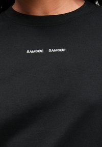 Black cotton t-shirt featuring two white embroidered "SAMSNØ" logos centered on the chest. Short sleeves and a round neckline.