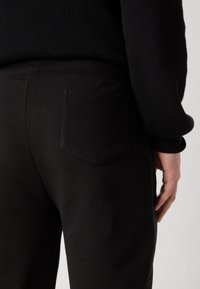 Persona vestida con suéter negro acanalado y pantalones negros con bolsillo trasero, de pie contra un fondo claro y liso, con la mano derecha visible.