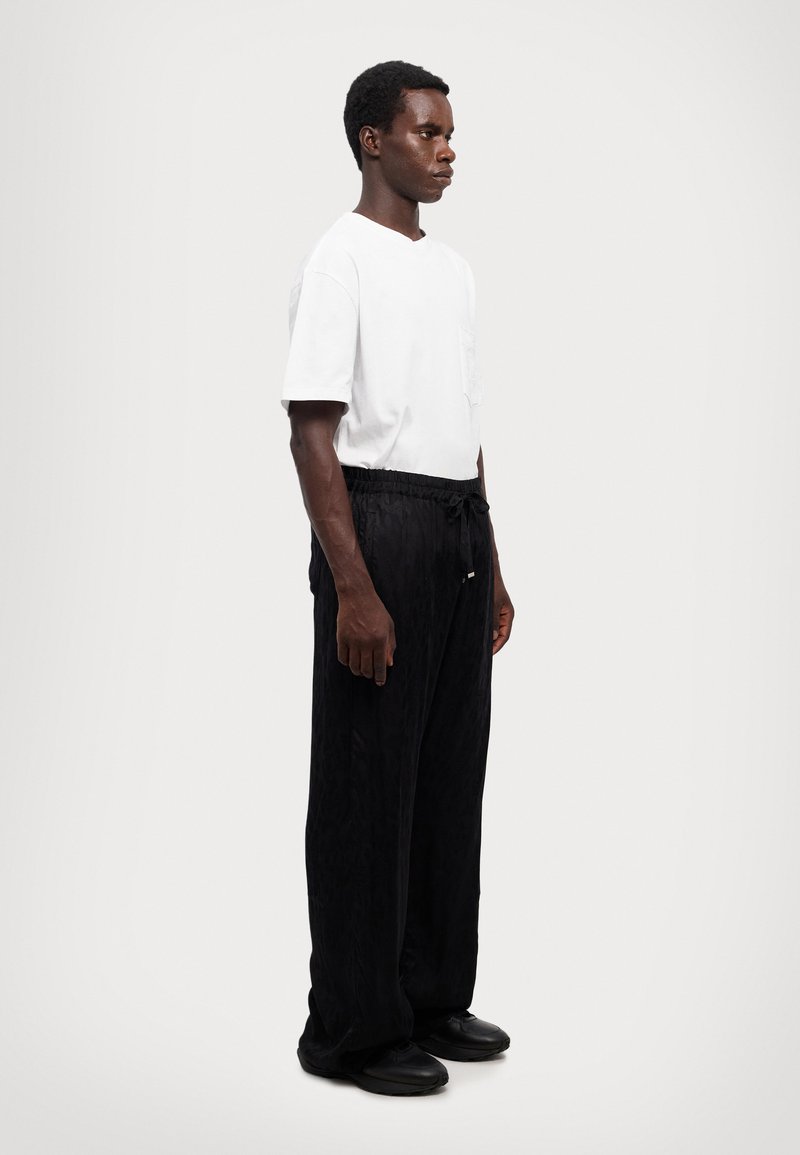 Vit kortärmad T-shirt, svarta byxor med dragsko i texturerat tyg och svarta slip-on skor. Modellen står framför en enfärgad bakgrund.