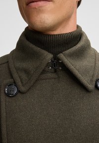 Manteau en laine vert foncé avec un col haut, arborant un tissage texturé, des accents de boutons noirs et une fermeture à clip au niveau du cou.
