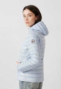 Lichtblauwe opgevulde jas met capuchon, diagonaal doorgestikt patroon, ritszakken en een logopatch op de linker mouw.